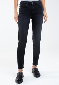 Jeans de mezclilla negra con un ajuste Slim, que cuentan con cierre de botón y diseño de cinco bolsillos. Textura suave y largo clásico hasta el tobillo. Zapatos negros sin cordones.