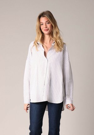 Chemise oversize blanche à rayures verticales bleues et rouges. Présente des manches longues, un col et une coupe décontractée. Jean en denim porté.