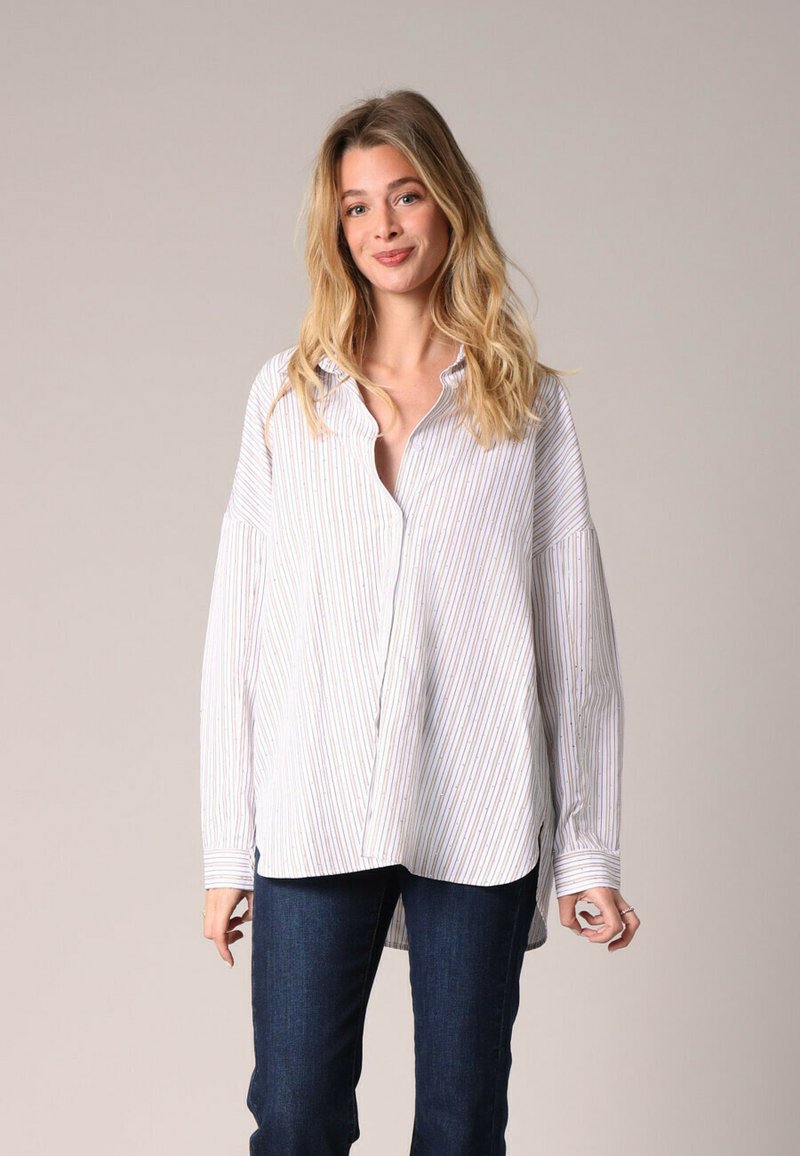 Oversized button-up shirt in white with vertical blue and red stripes. Features long sleeves, a collar, and a relaxed fit. Denim jeans worn.
