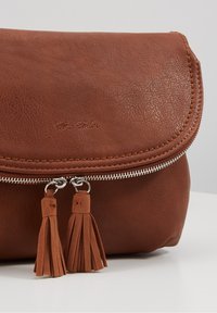 Brun läderclutch med en böjd flik, silverfärgad dragkedja och två dekorativa tofsar. Har en texturerad yta och präglat varumärkesnamn.