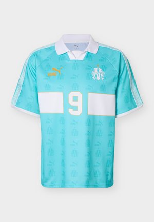 Ανοιχτό μπλε ποδοσφαιρική φανέλα με λευκό γιακά, μεγάλο λευκό αριθμό 9, κίτρινο λογότυπο Puma και έμβλημα του συλλόγου Μασσαλίας στο στήθος.