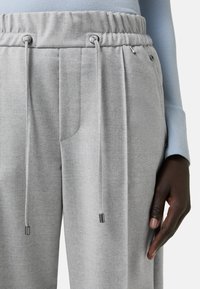 Pantalon de jogging gris avec une finition texturée, présentant une taille élastique, un cordon de serrage ajustable et une poche latérale. Tissu lisse avec un léger éclat.