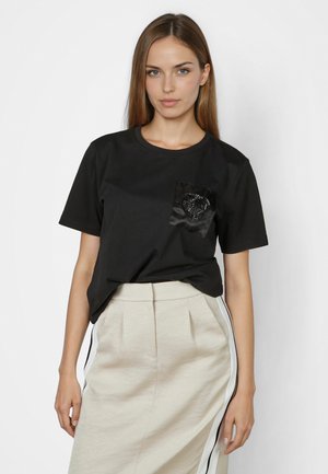 Young woman with straight brown hair wearing black short-sleeve shirt with beaded heart pocket and beige skirt with side stripes.