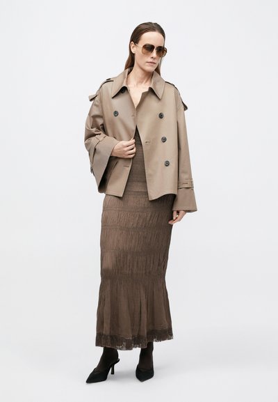 Beige dubbelknoop trenchcoat met brede mouwen over een gelaagde bruine rok met franjes, gecombineerd met zwarte, puntige hakken.