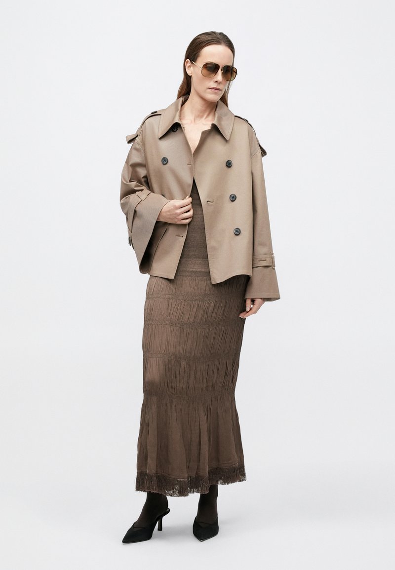 Trench-coat beige croisé à manches larges, porté sur une jupe marron à volants avec des détails en franges, accompagné de talons noirs pointus.