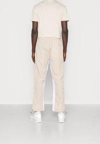 Beiga byxor med vita sidostråk, lättviktigt material, elastisk midja och dragkedjor vid anklarna, tillsammans med en ljusbeige t-shirt.