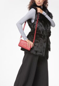 Gilet in finta pelliccia nera con vita cinturata, abbinato a pantaloni larghi a righe nere e borsa a tracolla rossa piccola in pelle.