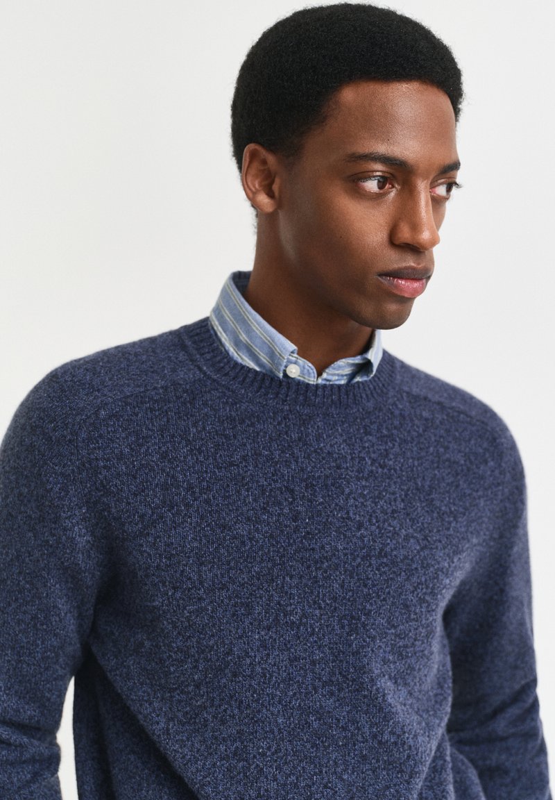 Giovane uomo con capelli corti e ricci che indossa un maglione blu girocollo sopra una camicia a righe abbottonata, che guarda verso destra su uno sfondo semplice.