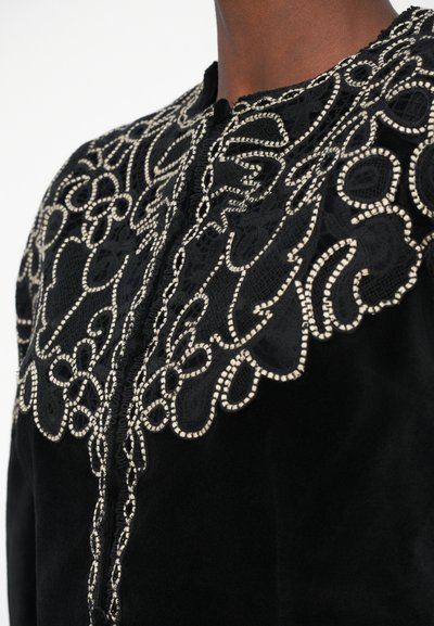Black velvet garment featuring intricate white embroidery along the neckline and front, with a lace-like design and button closure.