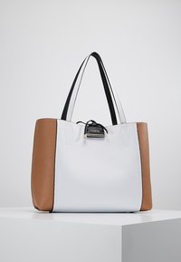 Sac cabas structuré bicolore avec devant blanc, côtés marron, poignées doubles noires et blanches, et une petite étiquette noire avec logo GUESS à l'avant.