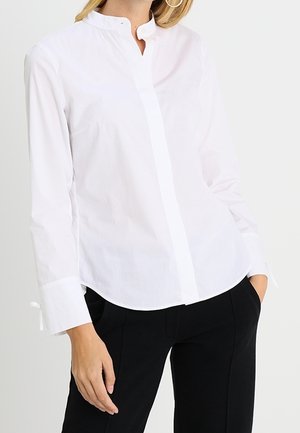Vrouw draagt een witte blouse met lange mouwen, een mandarijnkraag en een verborgen knopenlijst, gecombineerd met een zwarte broek.