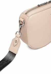 Béžová kožená crossbody taška zaobleného tvaru, se stříbrným zipem a vzorovaným černým popruhem připevněným kovovými prvky.