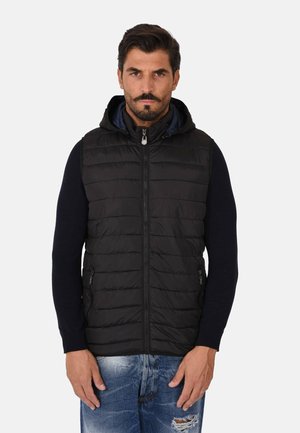 Gilet imbottito nero con cappuccio, dotato di zip sul davanti, cuciture verticali e tasche laterali. Indossato sopra una maglia scura a maniche lunghe.