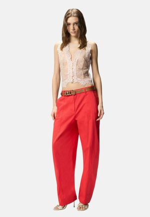 Giovane donna che indossa un top smanicato in pizzo trasparente, pantaloni rossi a gamba larga con cintura beige e tacchi aperti beige, in piedi davanti a uno sfondo semplice.