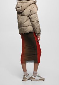 Chaqueta acolchada beige con capucha, usada sobre una falda de gradiente rojo y marrón hasta la rodilla, combinada con zapatillas blancas y grises.