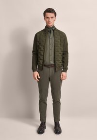 Olive green padded jacket with a high collar, buttoned shirt, and matching trousers. Dark brown shoes complete the outfit. Smooth texture throughout.