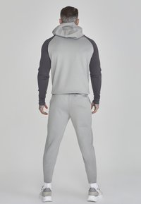 Tuta atletica grigia con cappuccio, caratterizzata da maniche scure a contrasto e una vestibilità affusolata. Il materiale appare liscio, con una combinazione di colori neutra.