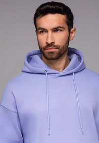 Sweatshirt violet avec capuche, arborant une coupe décontractée, des cordons de serrage et une texture douce. Des coutures sont visibles sur les manches.
