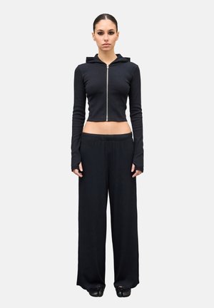 Mujer de pie mirando hacia adelante, vistiendo una sudadera negra corta con cremallera, pantalones anchos negros y zapatos negros sobre un fondo blanco.