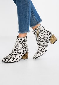 Białe botki za kostkę z czarnym plamkowanym wzorem. Złote akcenty na kwadratowym obcasie z ćwiekami, zamek błyskawiczny po boku oraz teksturowana powierzchnia.