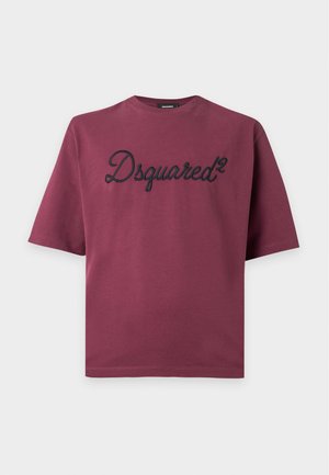 Junger Mann trägt ein dunkelrotes Dsquared2 T-Shirt, blaue ausgewaschene Jeans und beigefarbene Sneaker, steht vor einem schlichten hellen Hintergrund.