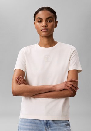 Γυναίκα με σκούρα πίσω μαλλιά, που φοράει λευκό κοντομάνικο πουκάμισο και ανοιχτό μπλε τζιν, στέκεται με σταυρωμένα χέρια μπροστά σε γκρίζο φόντο.