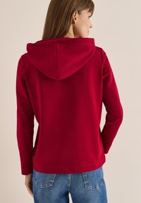Czerwona bluza z kapturem z regulowanym sznurkiem, długimi rękawami i luźnym krojem. Wykonana z miękkiego materiału, ma prosty dół. Połączona z niebieskimi dżinsami.