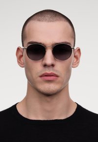 Ümar mustade prillide tume lense, õhukesed hõbedased metallraamid ja minimalistlik disain, kantud inimese poolt neutraalses taustas.