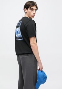 Czarny t-shirt z kolorową grafiką gór na plecach. Szare spodnie i niebieska czapka trzymana w prawej ręce. Prosty, codzienny design.