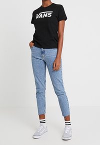 Camiseta de algodón negra con el logo "VANS", combinada con jeans de mezclilla azul claro con dobladillos deshilachados. Zapatillas blancas con detalles grises y calcetines a rayas.