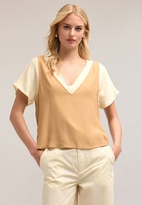 Top senza maniche con scollatura a V in beige e crema, caratterizzato da una texture liscia e una vestibilità comoda, abbinato a pantaloni chiari.