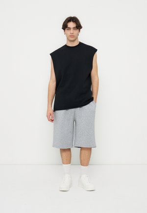 Jeune homme debout, portant un débardeur noir, un short gris aux genoux, des chaussettes blanches et des baskets blanches, sur fond blanc uni.