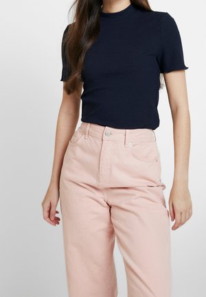 Kobieta w granatowym prążkowanym topie z krótkim rękawem, wpuszczonym w wysokowysokie, jasnoróżowe spodnie o szerokich nogawkach, na jednolitym tle.