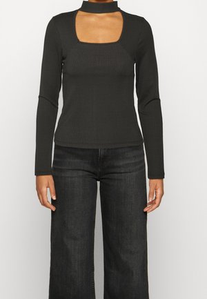Schwarzes, geripptes Langarmoberteil mit einem quadratischen Ausschnitt am Hals und einem hohen Kragen. Das figurbetonte Design kontrastiert mit lockeren, dunklen Jeans.