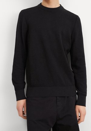 Person iført en ensfarvet sort langærmet sweater og sorte bukser, stående mod en hvid baggrund.