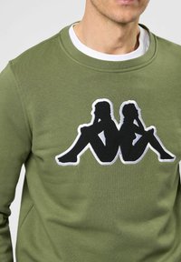 Olijfgroene sweatshirt met een zwart geborduurd ontwerp van twee zittende figuren omrand door wit. Ronde halslijn en zacht materiaal.