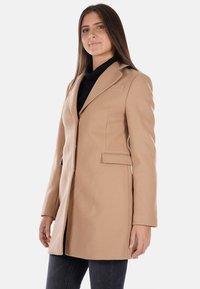 Cappotto beige su misura con revers, chiusura a un bottone e tasche laterali. Realizzato in un tessuto liscio con una vestibilità strutturata.