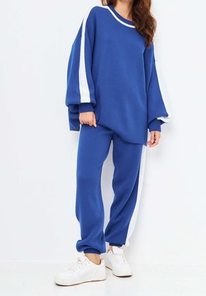 Femme portant un pull bleu oversize et un pantalon assorti avec des rayures blanches, associé à des baskets blanches, debout devant un fond blanc.