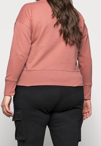 Sudadera rosa de manga larga con puños acanalados, que presenta un diseño recortado y un acabado suave y texturizado. Pantalones cargo negros visibles por debajo.