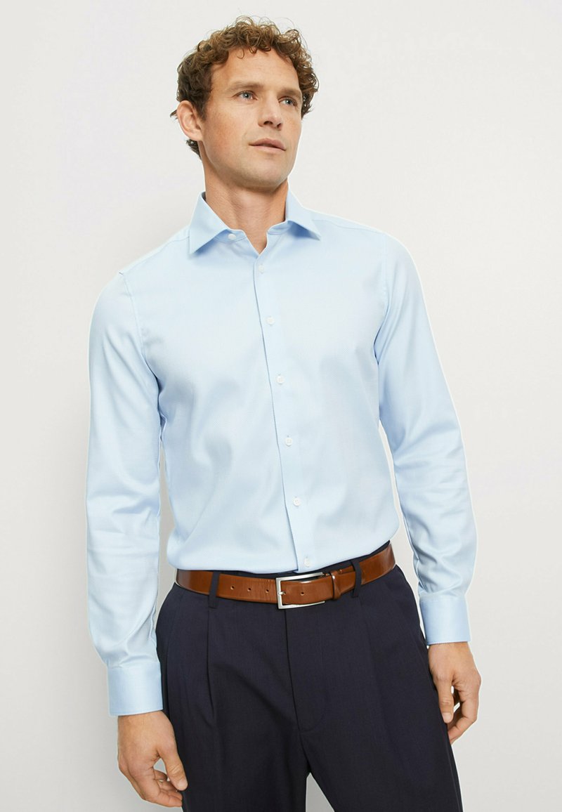 Hellblaues Hemd mit Knopfleiste aus strukturiertem Stoff, langen Ärmeln und weitem Kragen, kombiniert mit marineblauen Hosen und einem braunen Ledergürtel.