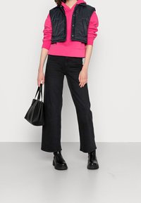 Rosa Kapuzenpullover, gesteppte schwarze Weste, schwarze weite Jeans und schwarze Knöchelstiefel. Hält eine kleine schwarze Handtasche.