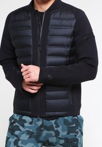 Černá bomber bunda s polstrovaným předním dílem, opatřená zipem a bočními kapsami. Hladká látka kontrastuje s prošívanou strukturou.