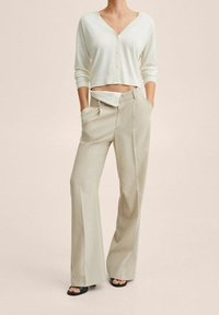 Frau trägt eine weiße, gekürzte V-Ausschnitt-Strickjacke mit Knöpfen, beigefarbene, weit geschnittene Hose mit offenem Reißverschluss, Hände in den Taschen und schwarze Sandalen mit offenem Zehenbereich.