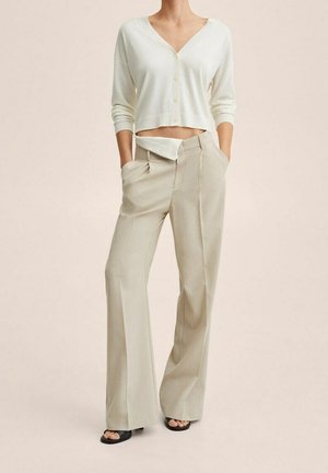 Nainen, joka pukeutuu valkoiseen, V-kaulaiseen lyhennettyyn neulejakkuun, beigenvärisiin leveälahkeisiin housuihin, joilla on avonaiset vetoketjut, kädet taskuissa, ja mustiin avoimiin sandaaleihin.