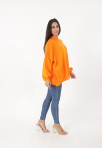 Übergroßer orangefarbener Strickpullover mit hohem Kragen und Ballonärmeln, kombiniert mit blauen Skinny-Jeans und transparenten High Heel-Sandalen.