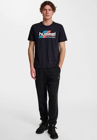 Czarny t-shirt Hummel z kolorowym logo, zestawiony z czarnymi spodniami dresowymi. Model ma na sobie czarne buty sportowe. Styl casual sportowy.