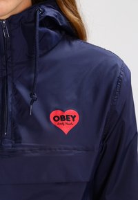 Navy windbreaker jacket with a half-zip, drawstring hood, and a red heart logo featuring "OBEY" and "lonely hearts" text. Smooth fabric.