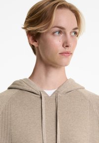 Beige hoodie with ribbed texture, drawstring, and hood. Features a white layered shirt underneath at the neck. Simple design, soft fabric.