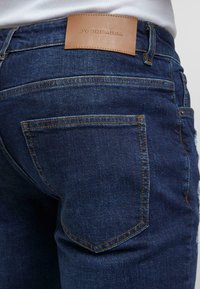 Mørkeblå jeans laget av denim med jevn tekstur, med et brunt lærpatch på midjen og kontrasterende gul søm.