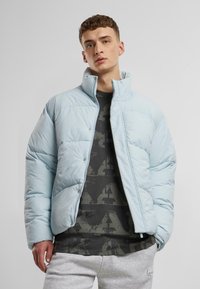 Ljusblå pufferjacka med hög krage, tryckknappar och kviltad textur, bärs över en mörkt mönstrad skjorta och grå sweatpants.